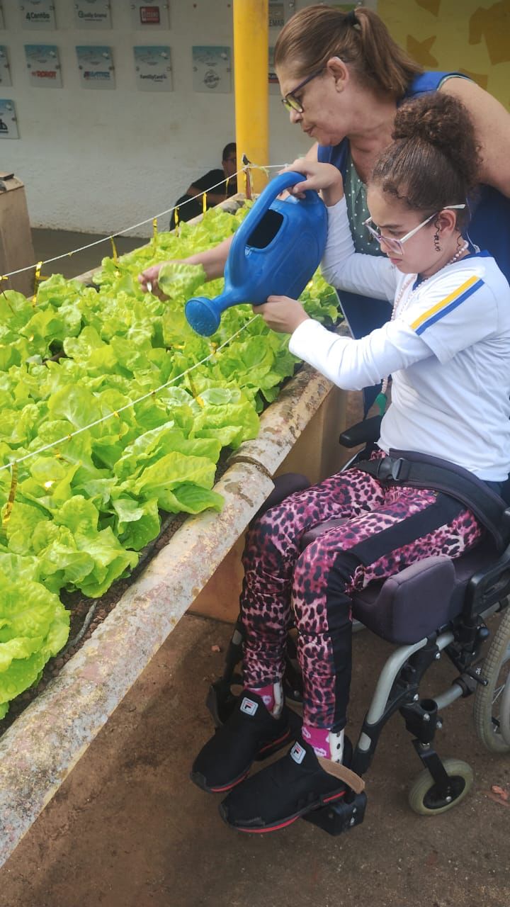 Aluna em cadeira de rodas regando a horta com apoio da educadora